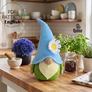 May include: A knitted gnome doll with a light blue pointed hat adorned with a white daisy. The gnome has a green body, a beige beard, and tan arms. The image includes the text "PDF PATTERN English".