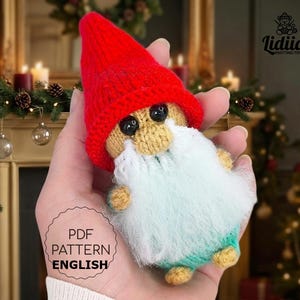 May include: A knitted gnome toy with a red hat, beige face, white beard, and blue-green body. The gnome has black eyes and is held in a hand. The image includes the text "PDF PATTERN ENGLISH".