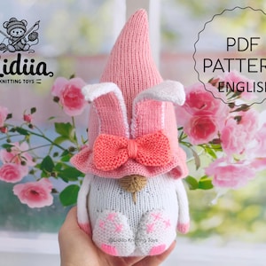 May include: A pink and white knitted bunny gnome with a pink bow. The gnome is holding a small white flower. The background is a blurry image of pink flowers.