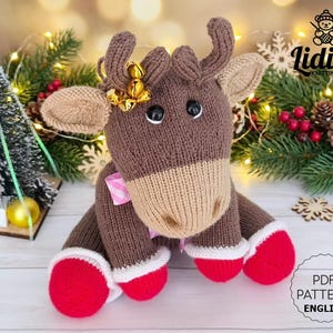 Puede incluir: Un juguete de reno tejido a mano en tonos marrón y beige, con detalles rojos y blancos en los pies. Tiene ojos negros, un lazo rosa y campanas doradas en sus cuernos. El juguete es un patrón PDF en inglés. Decoración navideña.