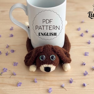 Könnte beinhalten: Ein weißer Keramikbecher mit dem Text PDF PATTERN ENGLISH in einem kreisförmigen Rahmen, der auf einem braunen, gestrickten Untersetzer in Hundeform steht. Der Hund hat Schlappohren, große Kunststoffaugen, eine cremefarbene Schnauze und eine schwarze Nase.