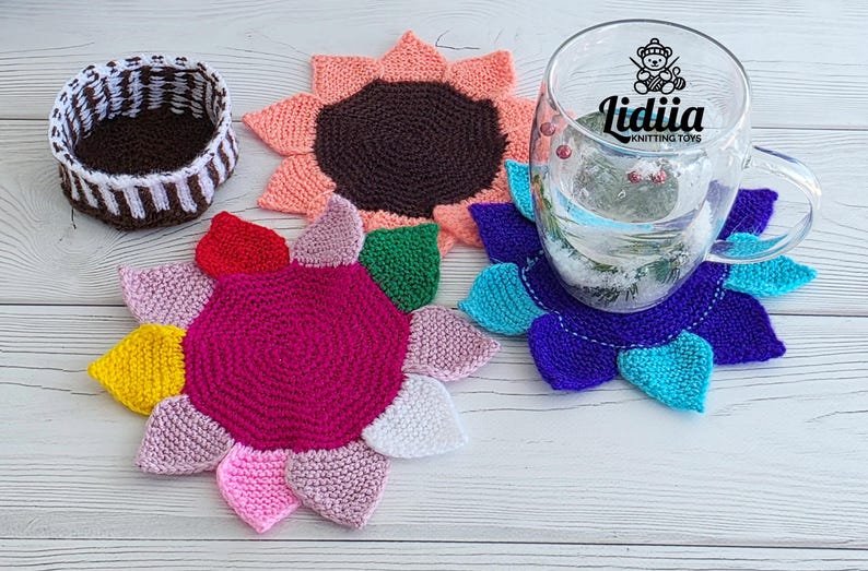Flat lay of three vibrant flower-shaped knitted coasters and a striped brown and white storage basket on a white wooden background.