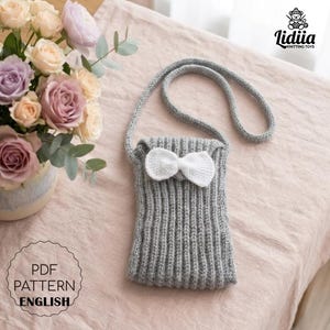 Puede incluir: Un bolso bandolera de punto gris con un lazo blanco. El bolso tiene una correa larga y está hecho de punto acanalado. La imagen también incluye un ramo de rosas y el texto "PDF PATTERN ENGLISH".