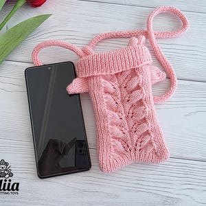 May include: A light pink knitted phone pouch with a long shoulder strap, featuring a vertical leaf-like cable knit pattern on the front and a folded top edge. A black smartphone is positioned next to the pouch for scale, with the Lidiia Knitting Toys logo.
