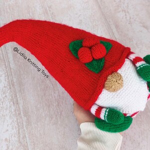 May include: A handmade Christmas gnome knitting pattern with a long, red knit hat and a white beard. The gnome has green mittens and a red and white striped body. The hat is adorned with green leaves and red berries. The text "@Lidiia Knitting Toys" is visible.
