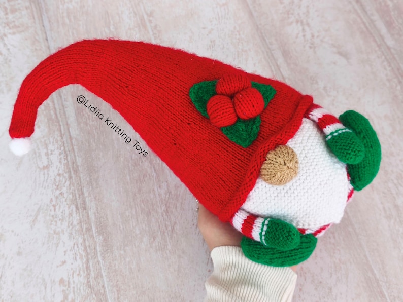 May include: A handmade Christmas gnome knitting pattern with a long, red knit hat and a white beard. The gnome has green mittens and a red and white striped body. The hat is adorned with green leaves and red berries. The text "@Lidiia Knitting Toys" is visible.