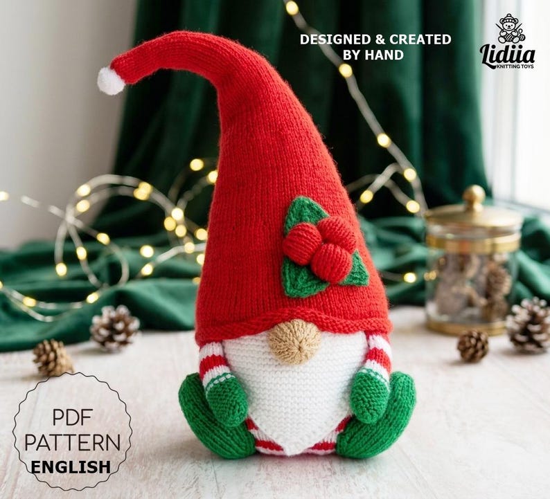 A knitted gnome toy featuring a tall red hat with a white pom-pom tip and a green holly leaf and red berry embellishment. The gnome has a white beard, a tan nose, and green mittens and boots with red and white striped sleeves and legs.