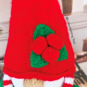 May include: A red knitted gnome hat featuring a decorative applique of three red berries and two green leaves. The text @Lidiia Knitting Toys is printed vertically on the right side of the red fabric. The gnome has a beige nose and striped sleeves.