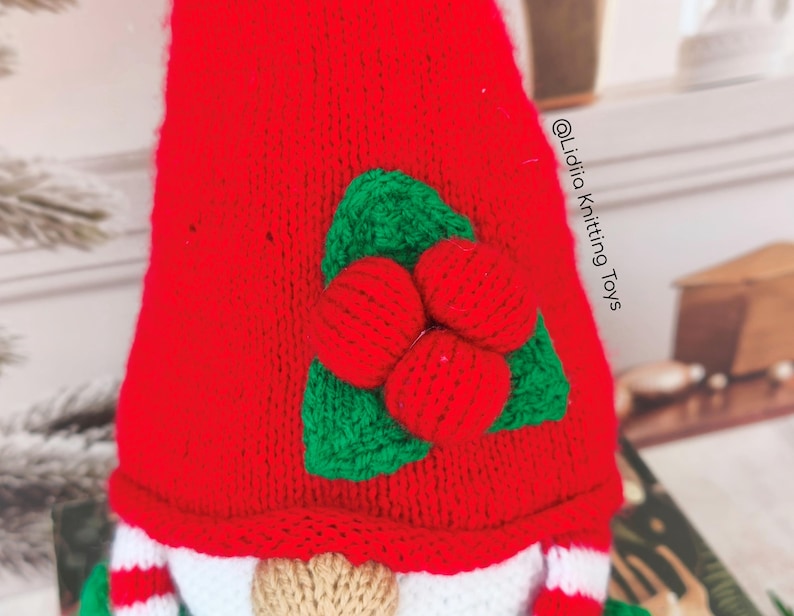 May include: A red knitted gnome hat featuring a decorative applique of three red berries and two green leaves. The text @Lidiia Knitting Toys is printed vertically on the right side of the red fabric. The gnome has a beige nose and striped sleeves.