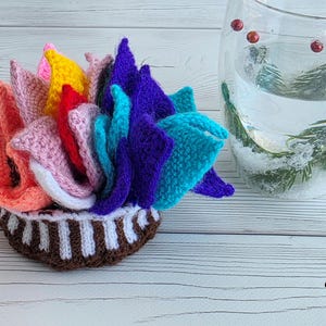 Brightly colored knitted flower mats folded together and stored inside a small handknit basket next to a glass cup.