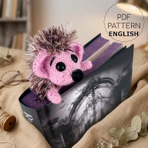 May include: A pink and brown knitted hedgehog bookmark peeking out from a book. The bookmark has black eyes and a black nose. The book has a dark cover with a grayscale image. Text on the image reads "PDF PATTERN ENGLISH".