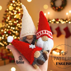 May include: Three knitted gnome dolls with red and cream-coloured hats, grey beards, and grey and brown bodies. One gnome has a snowflake on its hat. The gnomes are held in a hand, with a Christmas tree and fireplace in the background.