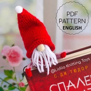 May include: A handmade red knitted gnome bookmark with a white beard and a small white pom-pom on the hat. The bookmark is holding a book with text in Russian. The image includes the text "PDF PATTERN ENGLISH".
