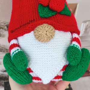 May include: A crocheted gnome doll featuring a bright red hat with a green leaf and red berry accent, a tan nose, a white beard, and green mittens. The gnome wears red and white striped sleeves and pants, with the text @Lidiia.Knitting.Toys visible.