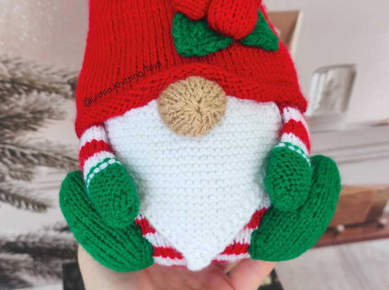 May include: A crocheted gnome doll featuring a bright red hat with a green leaf and red berry accent, a tan nose, a white beard, and green mittens. The gnome wears red and white striped sleeves and pants, with the text @Lidiia.Knitting.Toys visible.