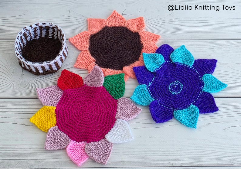 Flat lay of three vibrant flower-shaped knitted coasters and a striped brown and white storage basket on a white wooden background.