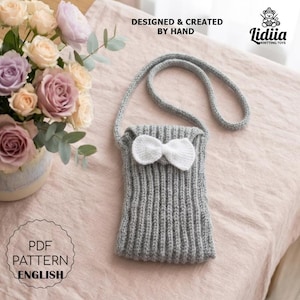 Può includere: Una borsa a tracolla lavorata a maglia grigia con una trama a coste e un fiocco all'uncinetto bianco sul davanti. La borsa include una tracolla lunga ed è accompagnata da un bouquet di rose rosa e color lavanda. Il testo recita DESIGNED & CREATED BY HAND, Lidiia KNITTING TOYS e PDF PATTERN ENGLISH.