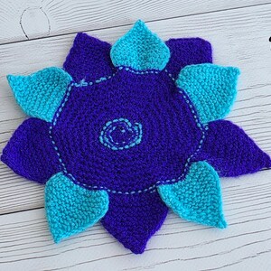 May include: A crocheted flower-shaped mat featuring a dark purple center with a light blue spiral detail at the middle. The mat has alternating dark purple and bright turquoise petals arranged in a star pattern with visible yarn texture.