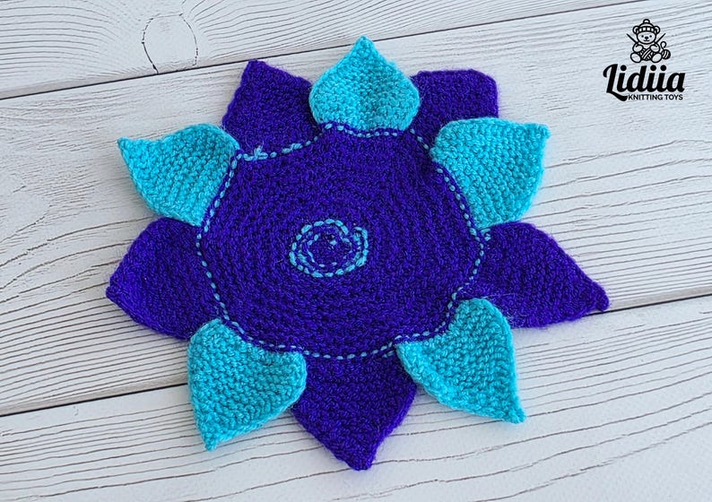 May include: A crocheted flower-shaped mat featuring a dark purple center with a light blue spiral detail at the middle. The mat has alternating dark purple and bright turquoise petals arranged in a star pattern with visible yarn texture.