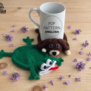 May include: A white ceramic mug with the text PDF PATTERN ENGLISH printed on the front. A brown knitted dog toy with black eyes and a white snout, and a green knitted frog toy with white eyes and a red tongue are positioned near the mug.