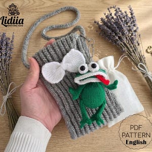 May include: A handmade gray knitted bag with a white bow and a green frog keychain. The frog has a red tongue and large eyes. The bag has a gray strap and is next to a bundle of lavender.