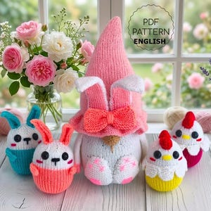 Puede incluir: Una colección de cinco figuras tejidas que incluye un gnomo grande con sombrero rosa y orejas de conejo, dos conejitos pequeños en azul y coral, y dos pollitos pequeños en amarillo y rosa. El texto PDF PATTERN ENGLISH es visible arriba a la derecha.