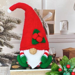 May include: A handmade knitted gnome featuring a tall, pointed red hat with a white pom-pom at the tip. The gnome has a beige nose, a white beard, green mittens, and red and white striped sleeves. A green leaf and red berry applique adorns the hat.