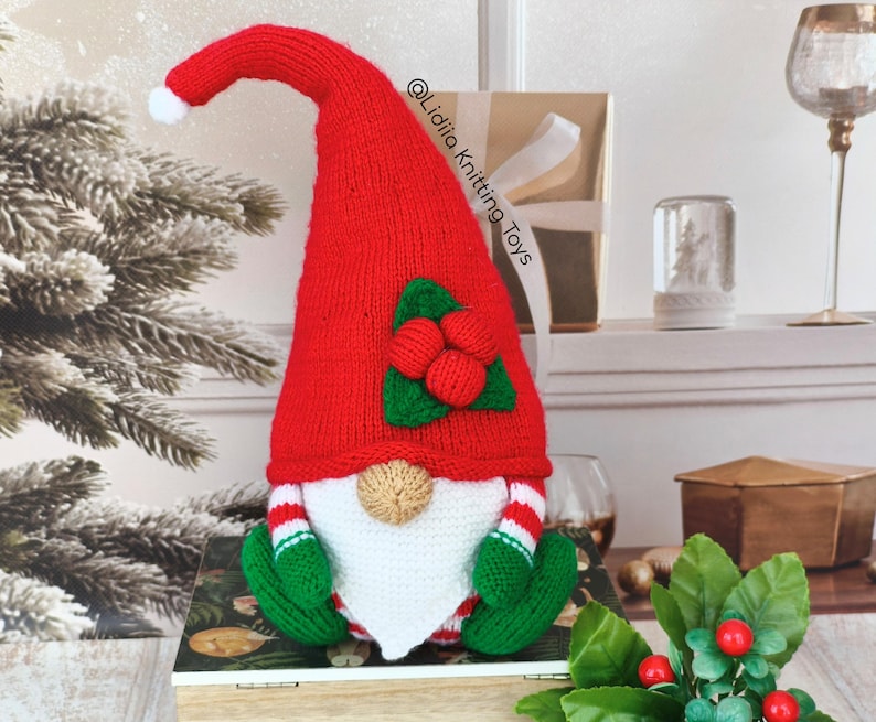 May include: A handmade knitted gnome featuring a tall, pointed red hat with a white pom-pom at the tip. The gnome has a beige nose, a white beard, green mittens, and red and white striped sleeves. A green leaf and red berry applique adorns the hat.