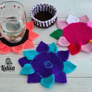Flat lay of three vibrant flower-shaped knitted coasters and a striped brown and white storage basket on a white wooden background.
