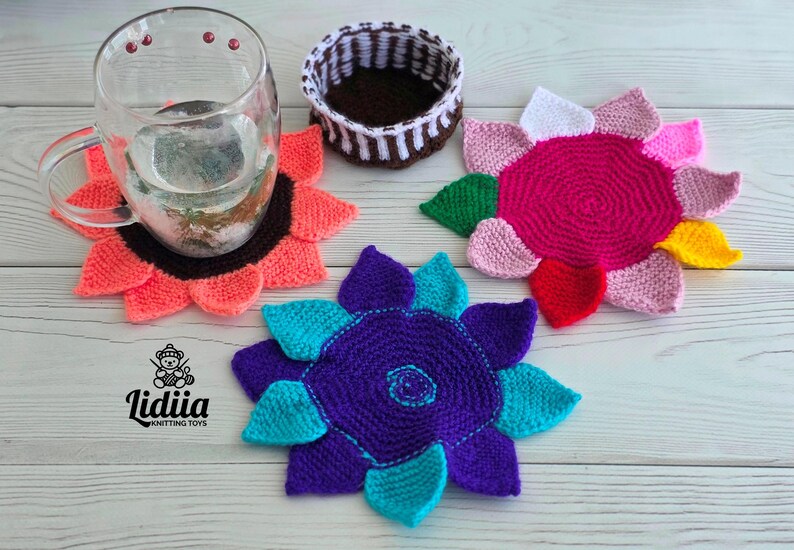 Flat lay of three vibrant flower-shaped knitted coasters and a striped brown and white storage basket on a white wooden background.