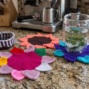 May include: A collection of three handmade knitted coasters shaped like flowers with multi-colored petals, including a pink, yellow, and white version, a coral and brown version, and a purple and teal version, alongside a small brown and white bowl.