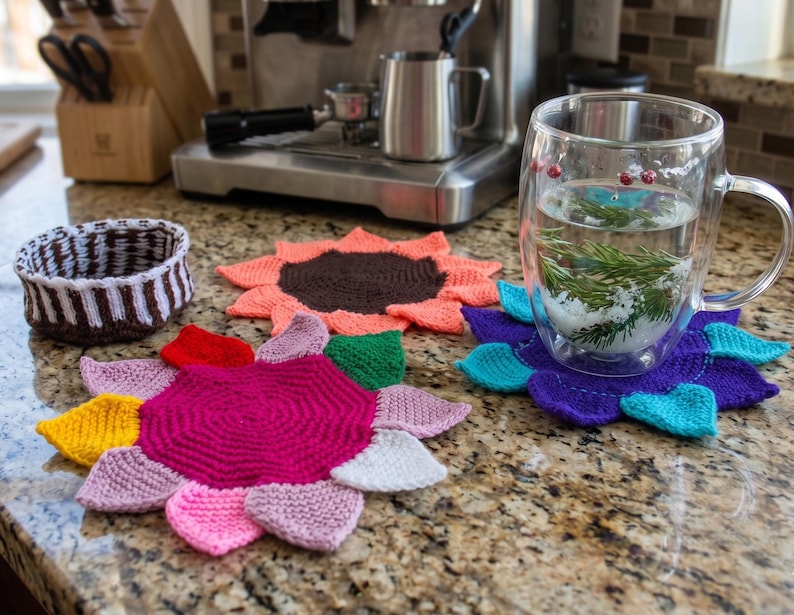 May include: A collection of three handmade knitted coasters shaped like flowers with multi-colored petals, including a pink, yellow, and white version, a coral and brown version, and a purple and teal version, alongside a small brown and white bowl.