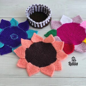 Flat lay of three vibrant flower-shaped knitted coasters and a striped brown and white storage basket on a white wooden background.