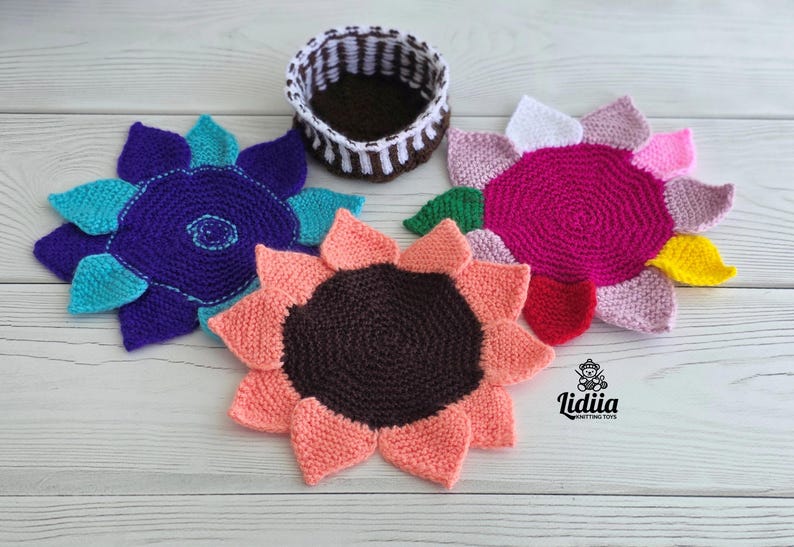 Flat lay of three vibrant flower-shaped knitted coasters and a striped brown and white storage basket on a white wooden background.