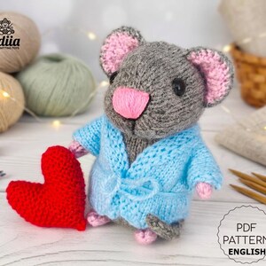 May include: A hand-knitted gray mouse toy with pink inner ears and a pink nose, wearing a light blue knitted robe with a tie waist. The mouse is holding a bright red knitted heart in its hand. The text Lidiia Knitting Toys is visible.