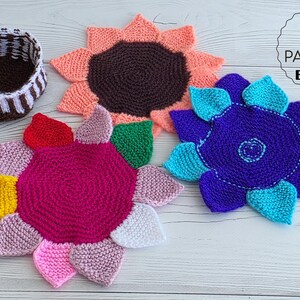 Flat lay of three vibrant flower-shaped knitted coasters and a striped brown and white storage basket on a white wooden background.