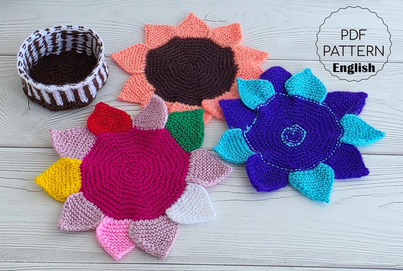 Flat lay of three vibrant flower-shaped knitted coasters and a striped brown and white storage basket on a white wooden background.