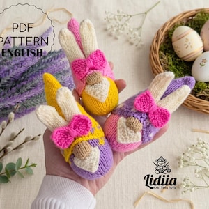 Pode incluir: Três brinquedos de coelhinho da Páscoa feitos à mão em tricô rosa, amarelo e roxo, cada um com um laço e um rosto creme. A imagem também mostra uma cesta com ovos de Páscoa decorados e o texto "PDF PATTERN ENGLISH".
