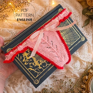 Op de afbeelding: Een roze gebreide boekenlegger met een rode rand en een strik, rustend op een donkerblauw boek met gouden details. De tekst "PDF PATTERN ENGLISH" is zichtbaar. De scène is geplaatst op een kanten oppervlak met bloemmotieven.