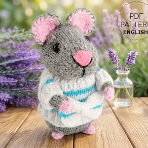 May include: A handmade knitted mouse toy in gray, white, and pink, wearing a sweater with blue stripes. The mouse has pink ears, a pink nose, and black eyes. The image includes the text "PDF PATTERN ENGLISH".