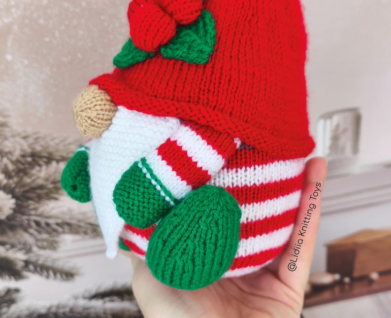 May include: A knitted gnome toy featuring a red hat with a green leaf and berry accent, a beige nose, a white beard, and a body with red and white horizontal stripes. The gnome has green arms and feet, with the text Lidiia Knitting Toys visible.