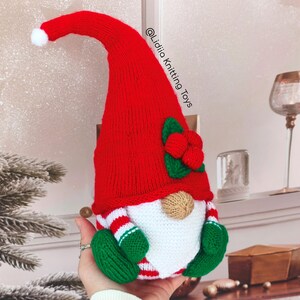 May include: A handmade knitted gnome toy featuring a tall red hat with a white pom-pom, a beige nose, a white beard, and green mittens. The gnome has red and white striped sleeves and the text @Lidiia Knitting Toys is visible on the hat.