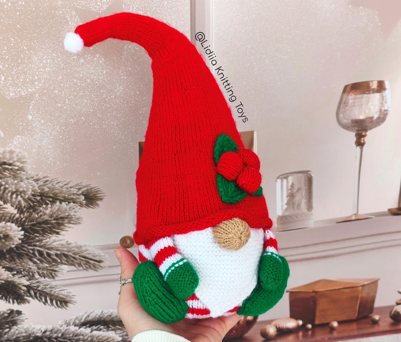 May include: A handmade knitted gnome toy featuring a tall red hat with a white pom-pom, a beige nose, a white beard, and green mittens. The gnome has red and white striped sleeves and the text @Lidiia Knitting Toys is visible on the hat.