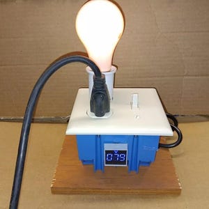 Puede incluir: Una toma de corriente blanca con una bombilla enchufada y un voltímetro digital que muestra 0,79 voltios. La toma de corriente está montada sobre una base de madera.