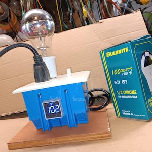 Op de afbeelding: Een blauwe stopcontact met een digitale voltmeter die 102 aangeeft, een heldere gloeilamp en een zwart elektriciteitskoord. Het stopcontact is verbonden met een witte elektrische doos. Een groene en gele doos met de tekst "BULBRITE. 100WATT 120V A-21 (5") 1/2 CHROME TOP MIRRORED BULB" bevindt zich op de achtergrond.