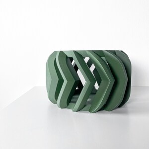 Könnte beinhalten: Ein grüner geometrischer Pflanzer mit einem einzigartigen, offenen, gewebten Design. Der Pflanzer ist aus einem glatten, festen Material gefertigt.