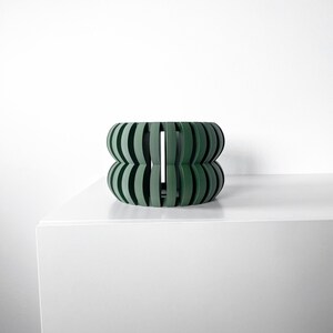 Könnte beinhalten: Ein dunkelgrüner, geometrischer, zylindrischer Pflanzer mit einem einzigartigen, offenen Design. Der Pflanzer ist aus einem glatten, harten Material gefertigt und hat eine moderne, minimalistische Ästhetik.