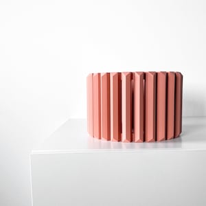 Könnte beinhalten: Ein geometrischer Pflanzer in Korallenfarbe aus Keramik. Der Pflanzer hat die Form eines Zylinders mit einer Reihe vertikaler, rechteckiger Öffnungen.
