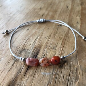 Könnte beinhalten: Ein schlichtes Armband mit drei Karneol-Steinen und silbernen Perlen auf einer weißen Schnur.