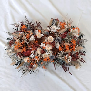 May include: A symmetrical floral arrangement of dried flowers in shades of orange, white, brown, and burgundy. The arrangement features a variety of textures and shapes, creating a visually appealing display. The background is a white fabric.